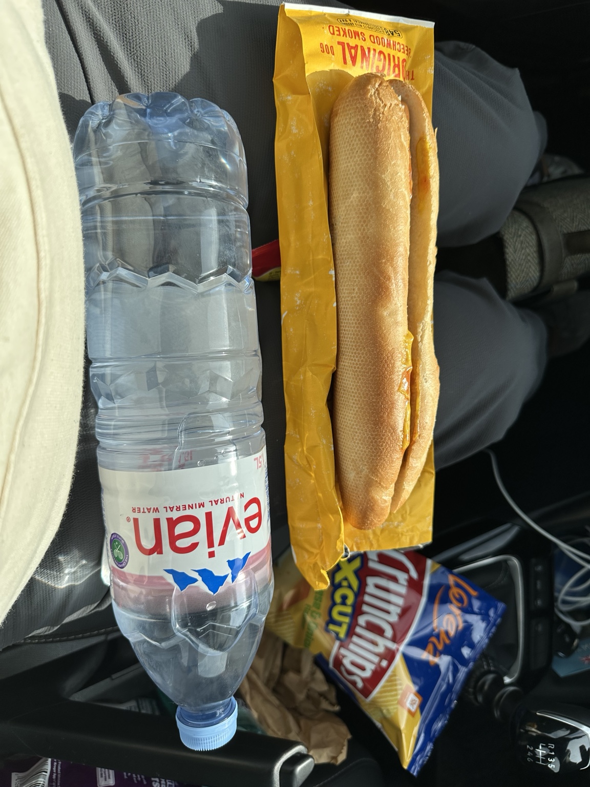 A very long hot dog beside water and crisps in the rental car